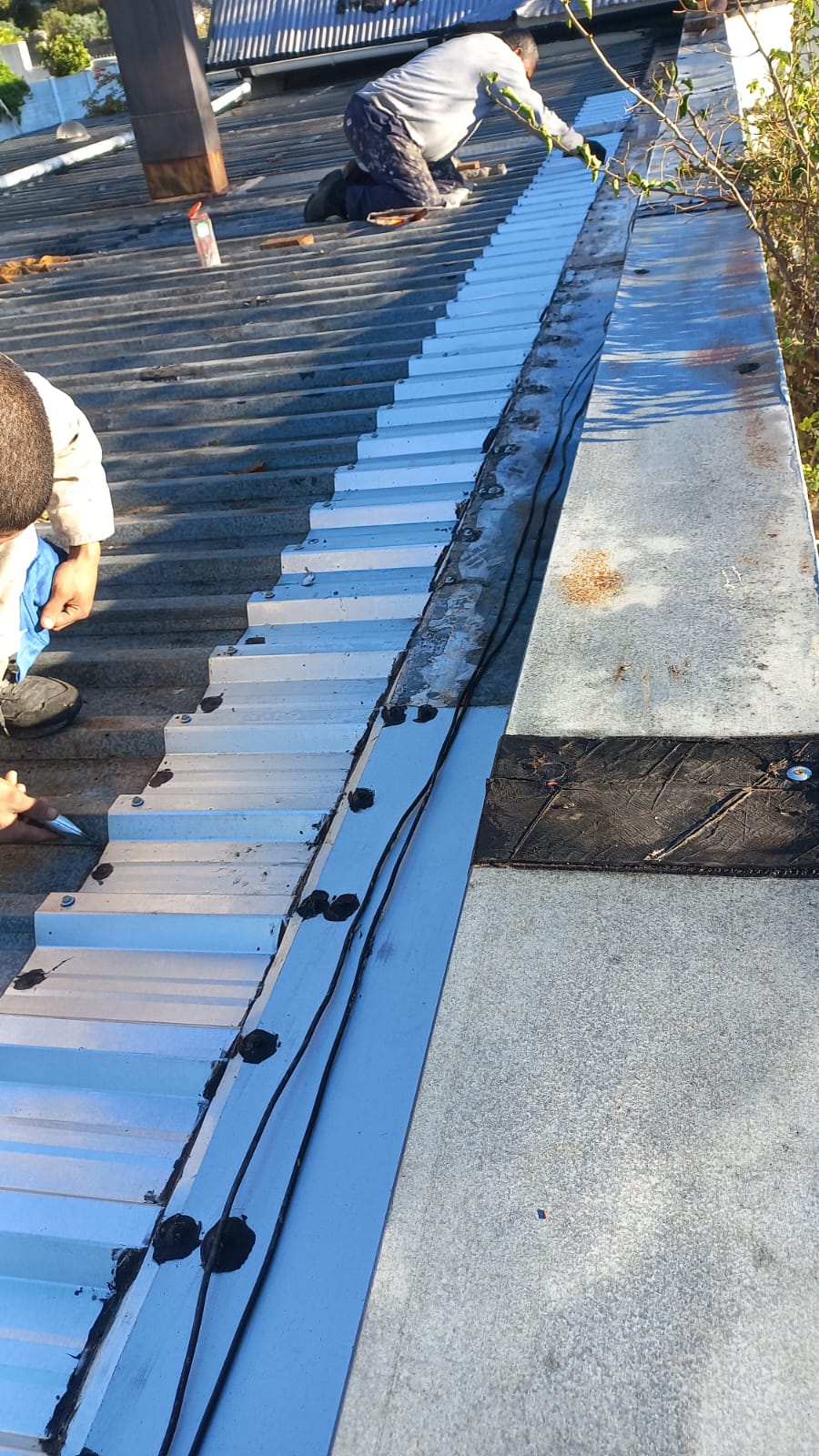 Workers repairing roof sheets and sealing a long strip beside the roof edge